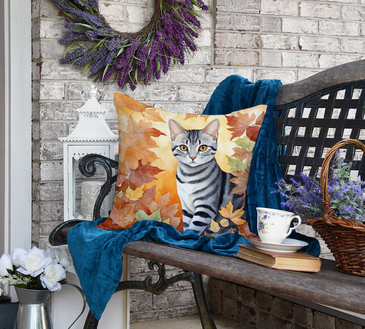 A decorated bench with a cat-themed autumn-leaf pillow, a blue blanket, a teacup on a book, a basket of lavender, and white roses in a metal pitcher.